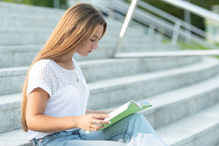 Pretty student with long blonde hair sitting on stairs reading a book.