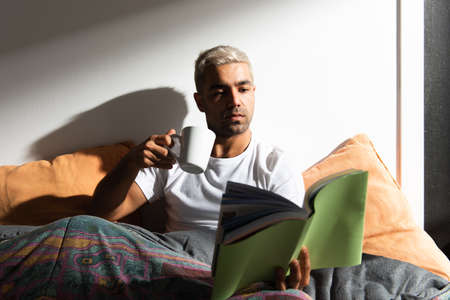 Hispanic man reading a book and drinking a cup of coffee on his bedの写真素材