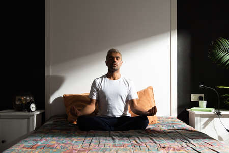 Hispanic man meditating in bedroom illuminated by sunlight. Healthy morning routine conceptの写真素材