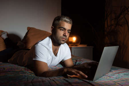 Hispanic man laying on bed and working on a laptop late at night.の写真素材
