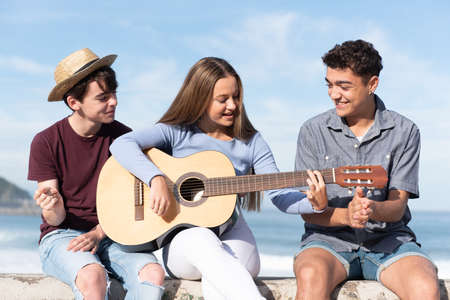 Group of happy multiethnic teenagers singing and playing the guitar sitting at the seaside.の写真素材