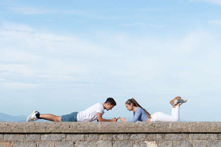 Teenager couple lying on a stone wall and using their phones. Digital addiction and dopamine concept. Copy space in the sky.の写真素材