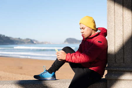 Latin man concentrated on his phone while sitting at seaside. Digital addiction concept.