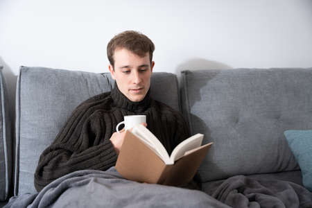Young man reading a book and taking a cup of coffee while sitting on sofa.