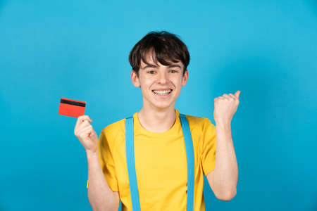 Teenager boy celebrating online purchase and holding credit card on blue background.