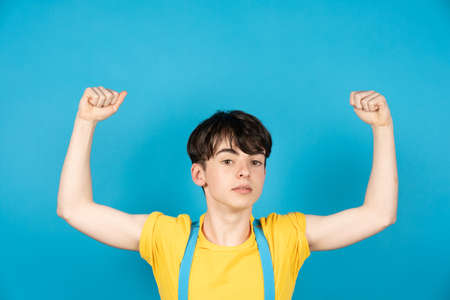 Slim teenager boy showing his biceps muscles. Arrogant adolescent on blue background. Flag colors of Ukraineの写真素材