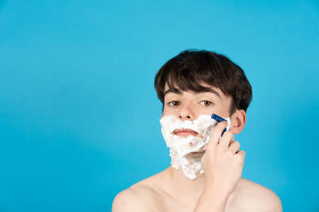 Portrait of teenager boy shaving face isolated on blue background.の写真素材
