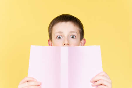 Surprised boy behind a book looking straight to camera with open eyes isolated on yellow background