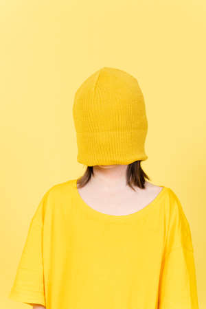 Portrait of blindfolded little girl wearing a big size t-shirt and wool hat isolated on yellow...