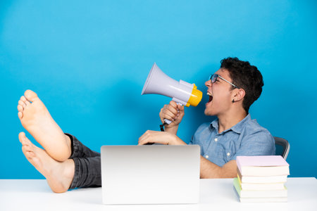 Hispanic teenager sitting feet on desk shouting on megaphone or speaker in front of laptop isolated on blue background.の写真素材