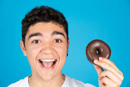 Happy hispanic teenager boy holding chocolate donut or donut and smiling to camera isolated on blue background.の写真素材