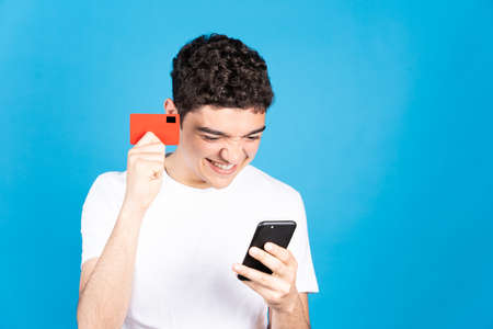 Hispanic teenager boy holding credit card and mobile phone isolated on blue background. Online...