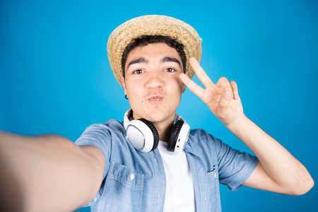 Hispanic young man making peace sign while taking a selfie isolated on blue background.の写真素材