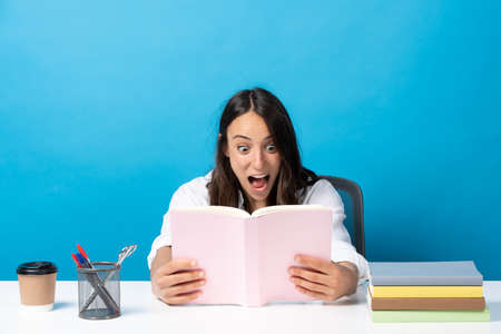 Surprised hispanic woman looking at book sitting behind desk isolated on blue background.