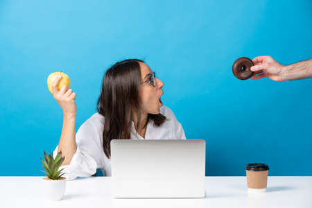 Hand offering donut to hispanic young woman holding fruit isolated on blue background. Food temptation in the office conceptの写真素材