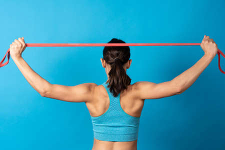 Back view of sportive hispanic woman using pilates rubber gum isolated on blue background. Muscle...