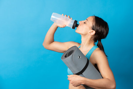 Hispanic sportswoman holding yoga mat and drinking water isolated on blue background.の写真素材