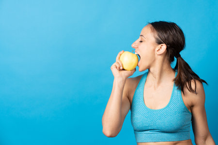 Hispanic sportswoman biting an apple with copy space isolated on blue background.