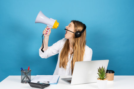 Angry telemarketing worker shouting on megaphone. Pretty displeased virtual assistance operator isolated on blue background.の写真素材