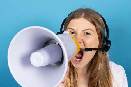 Closeup of virtual assistance operator shouting on megaphone isolated on blue background. Pretty telemarketing worker wearing headsetの写真素材