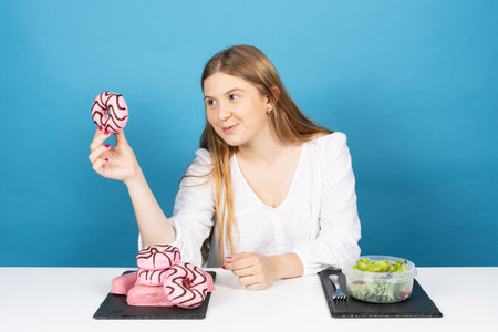 Young blonde woman sitting behind desk and holding donut isolated on blue background.. Salad and chocolate candy on table.の写真素材
