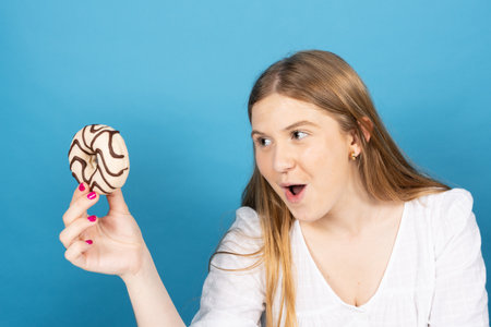 Pretty blonde teenager girl holding chocolate donut with envy isolated on blue background. Bad habits and temptation concept.の写真素材
