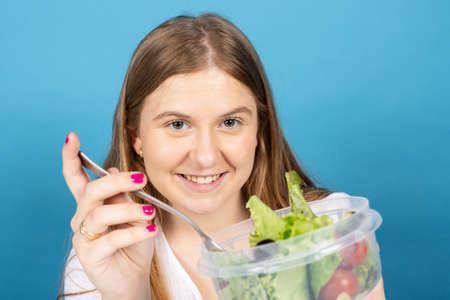 Portrait of chubby pretty woman eating salad and looking at camera isolated on blue background. Blonde teenager girl holding vegan food.の写真素材