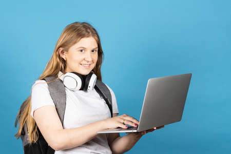 Pretty long haired college student typing on laptop. Blonde teenager girl isolated on blue...