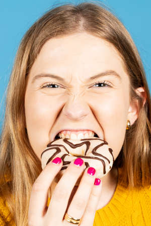 Blonde pretty teenager girl biting chocolate donut isolated on blue background. Bad eating habits...