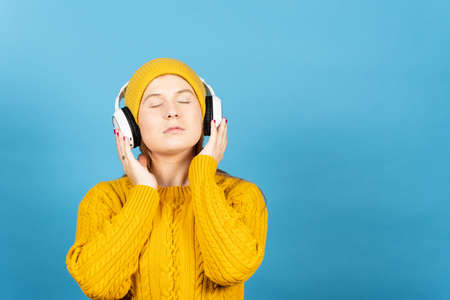 Winter clothing cute young woman listening relaxing music isolated on blue background. Teenager girl...