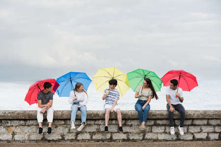 Group of friends talking sitting on a wall and holding colorful umbrellas