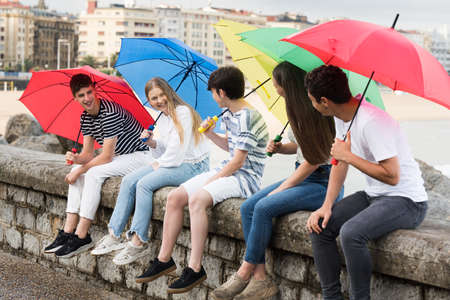 Group of teenagers talking while holding colorful umbrellas. Friends sitting on a wallの写真素材