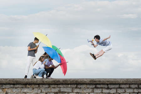 Abstract concept of ignoring toxic comments. Teenager boy shouting on megaphone and friends protecting themselves using umbrellas.の写真素材