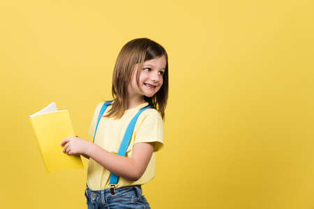 Cheerful little girl holding book and looking back at yellow copy space.の写真素材