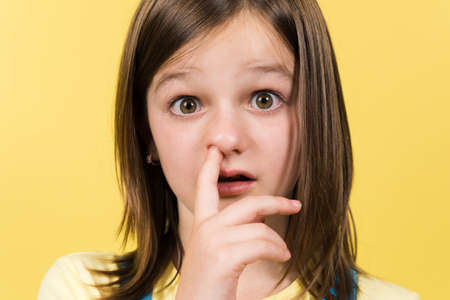 Little girl drilling the nose with finger to remove mucus isolated on yellow background. Funny kid portrait.の写真素材