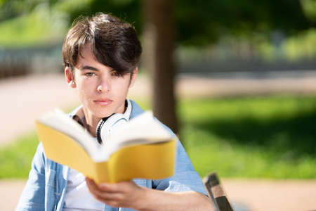 Closeup portrait of cute student boy looking at camera and holding book in a parkの写真素材
