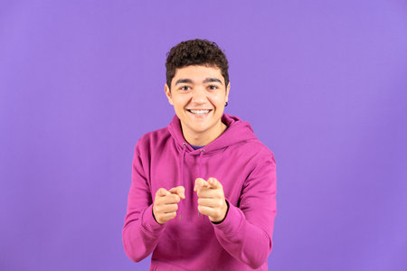Positive hispanic teenager boy smiling and pointing at camera isolated on purple backgroundの写真素材