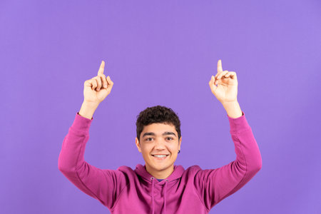 Smiling hispanic teenager boy pointing up at copy space isolated on purple background