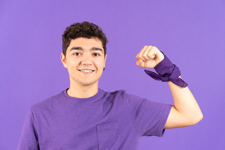 Front view of hispanic feminist teenager boy showing strength with raised arm isolated on purple...