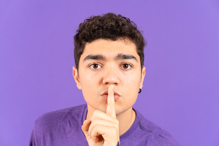 Closeup portrait of hispanic teenager boy with finger on mouth asking for silence isolated on purple backgroundの写真素材