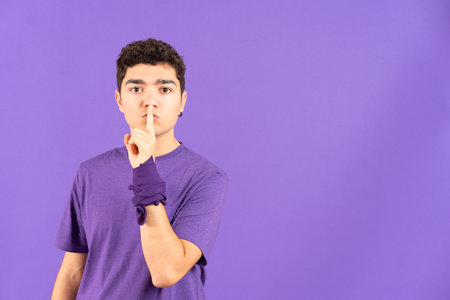 Hispanic teenager boy asking for silence finger on mouth isolated on purple background with copy space. Feminism and gender violence conceptの写真素材