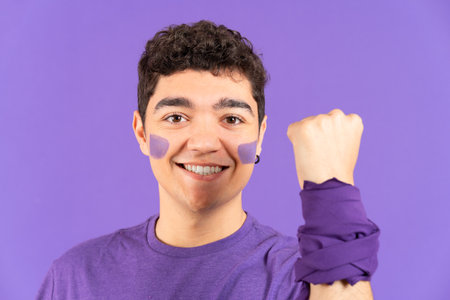 Smiling hispanic teenager boy with painted face and clenched fist isolated on purple background. Feminism conceptの写真素材