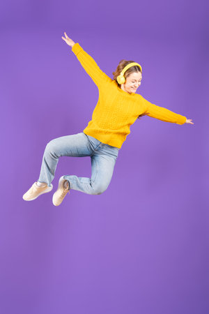 Pretty young woman dancing or levitating while listening to music on headphones isolated on purple...