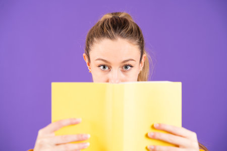 Pretty teenager girl looking at camera above book isolated on purple backgroundの写真素材