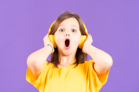 Surprised cute little girl listening to music or hearing podcast on yellow headphones isolated on purple backgroundの写真素材
