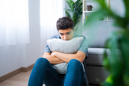 Sad hispanic teenager boy sitting on floor and hugging pillow. Depression and mental health concept.の写真素材