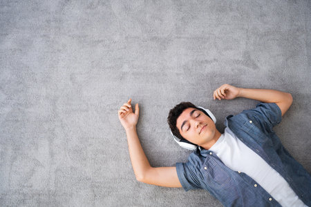 View from above of hispanic teenager boy listening to relaxing music on headphones and lying on carpet with copy space.の写真素材