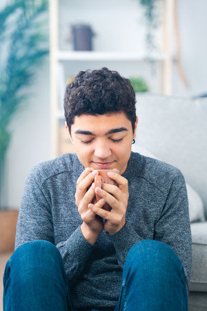 Hispanic teenager boy sitting on floor smelling cup of coffee.