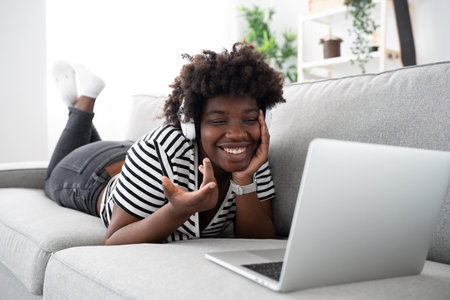 Happy young African American woman having video call on laptop while lying down on sofa. Black woman...