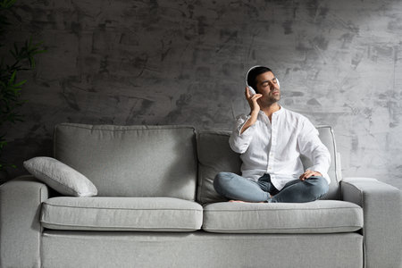 Hispanic young man listening relaxing music on headphones while sitting on sofa.の写真素材